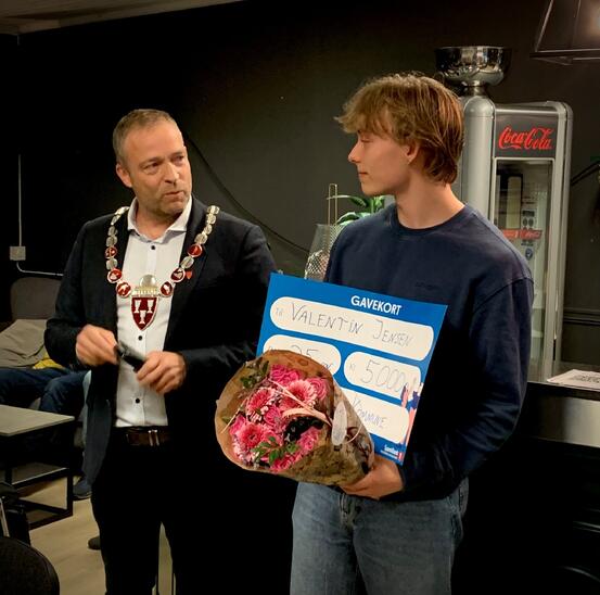 Bildet viser to personer. Den ene er ikledd ordførerkjede, den andre er yngre og står med blomster og en sjekk.