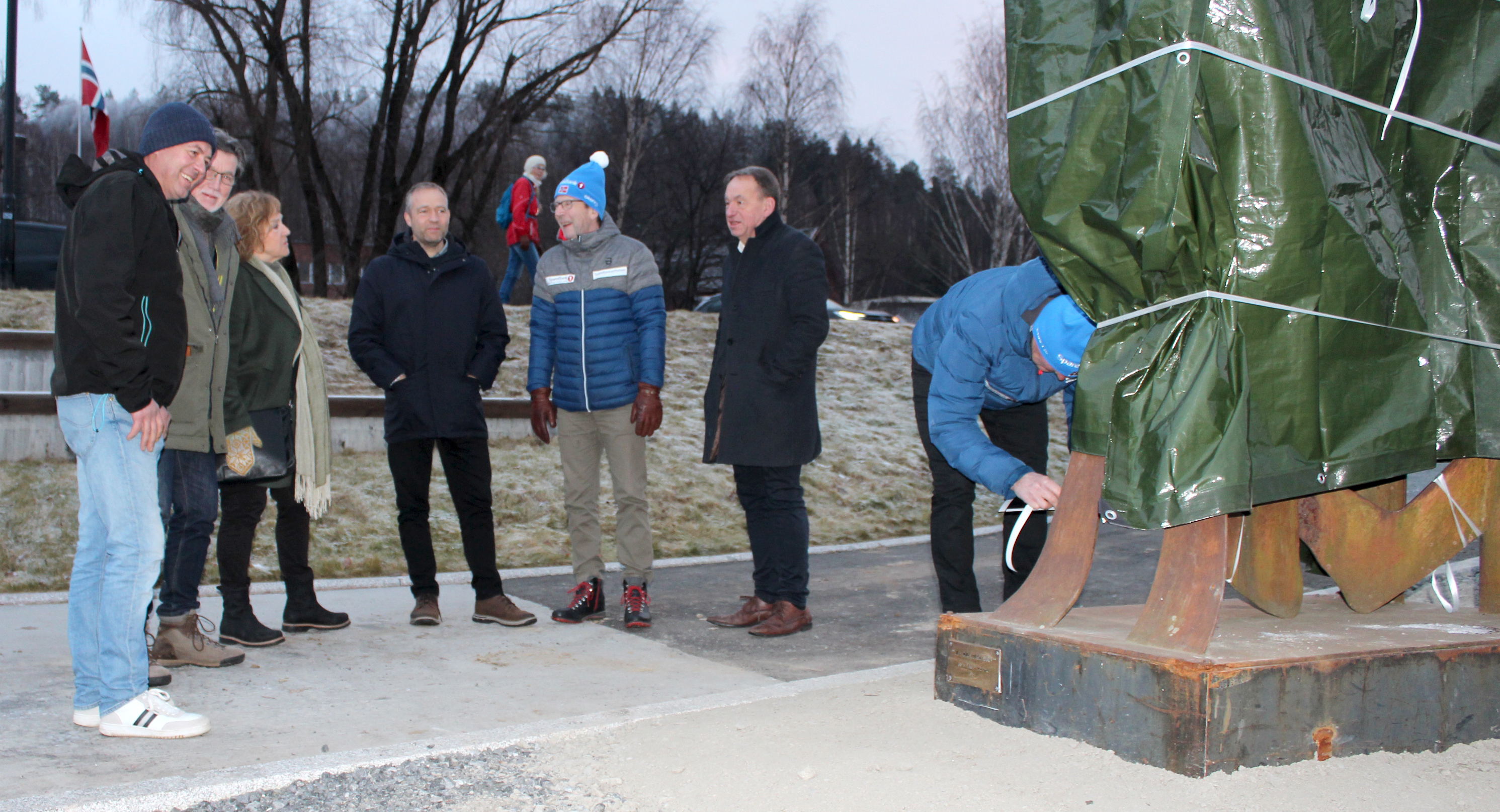 Bildet viser seks personer som står og prater sammen. En sjuende står bøyd og knyter opp en knute på et tau som holder fast en presenning. Denne er nærmest fotografen.