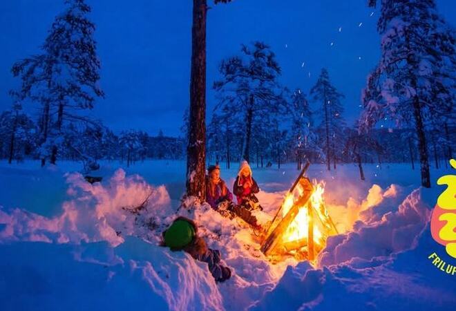 Bildet er et stemningsbilde med bål i snø, det er dunkelt. Det er en logo hvor det står «Friluftslivets år 2025».