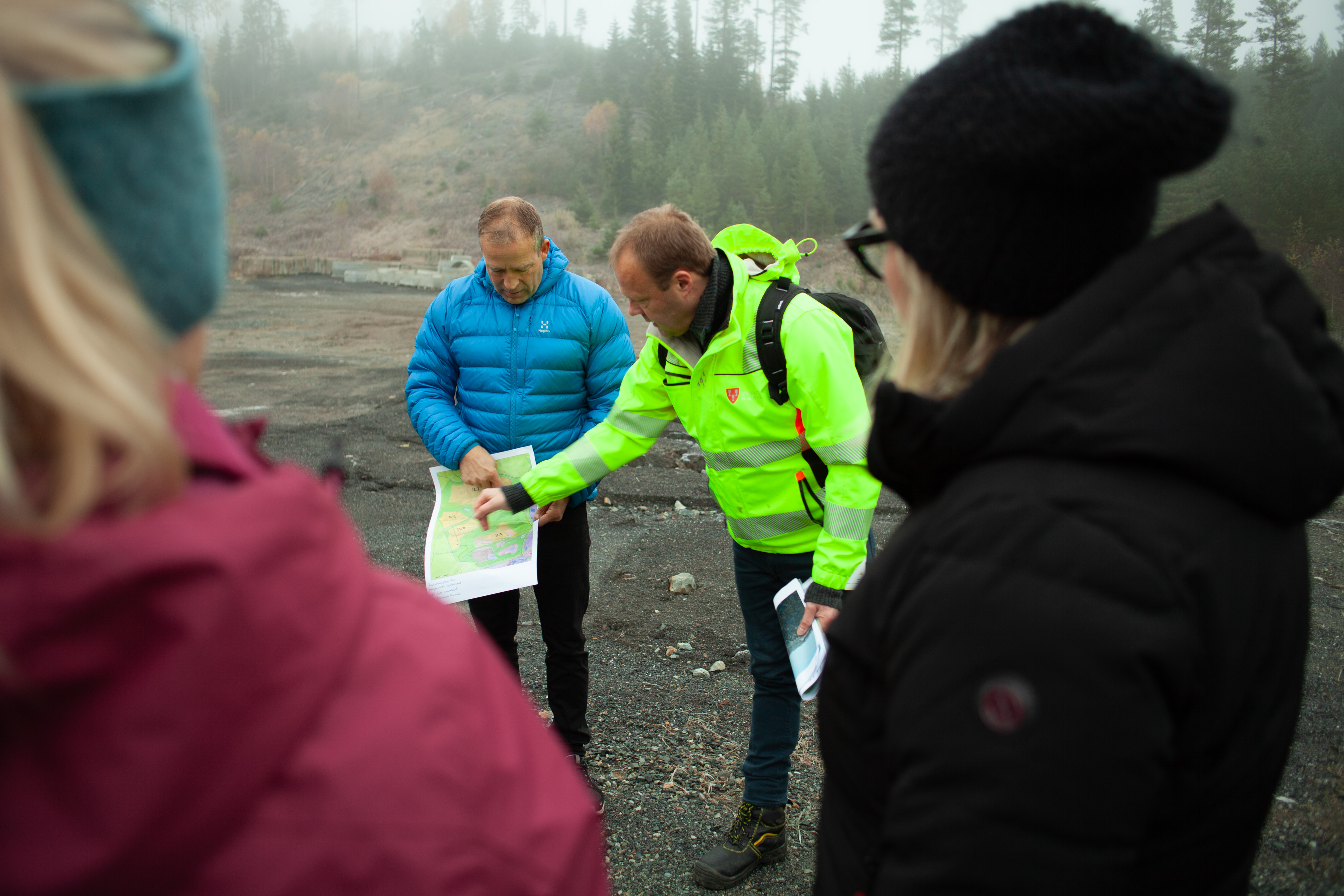 Bildet viser to personer som peker på et kart. Man ser ryggen til et par andre personer. I bakgrunnen ser man terreng.