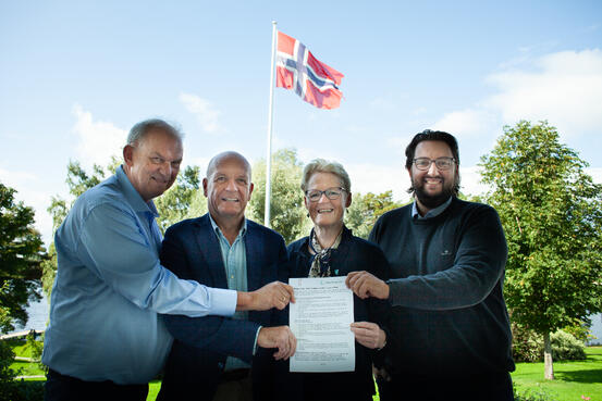 Bildet viser fire personer som står ved siden av hverandre. Alle holder i et papirark. De står ute. Det vaier et flagg bak dem.
