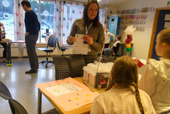 Kvinne står foran et bord med en ide og snakker med to elever