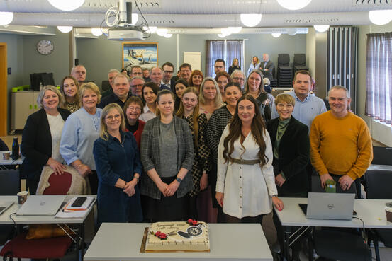 En stor gruppe mennesker står og smiler foran en kake.