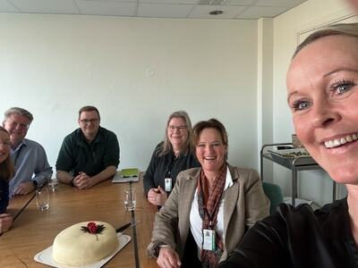 Bildet viser totalt seks personer. Fem sitter rundt et bord, i bakgrunnen for den som tar selfie. På bordet står en kake.
