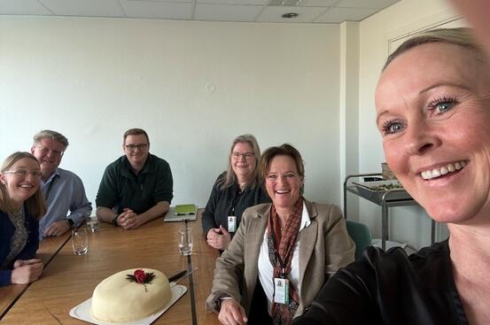 Bildet viser totalt seks personer. Fem sitter rundt et bord, i bakgrunnen for den som tar selfie. På bordet står en kake.