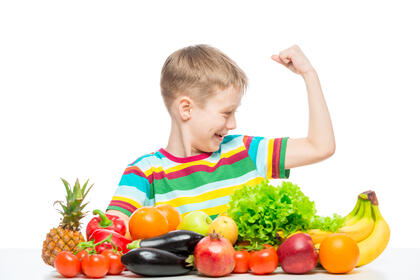 Bildet viser et barn som strammer musklene i overarmen, og ser på dem med et fornøyd smil. I forkant bugner det av fargerike grønnsaker og frukt.
