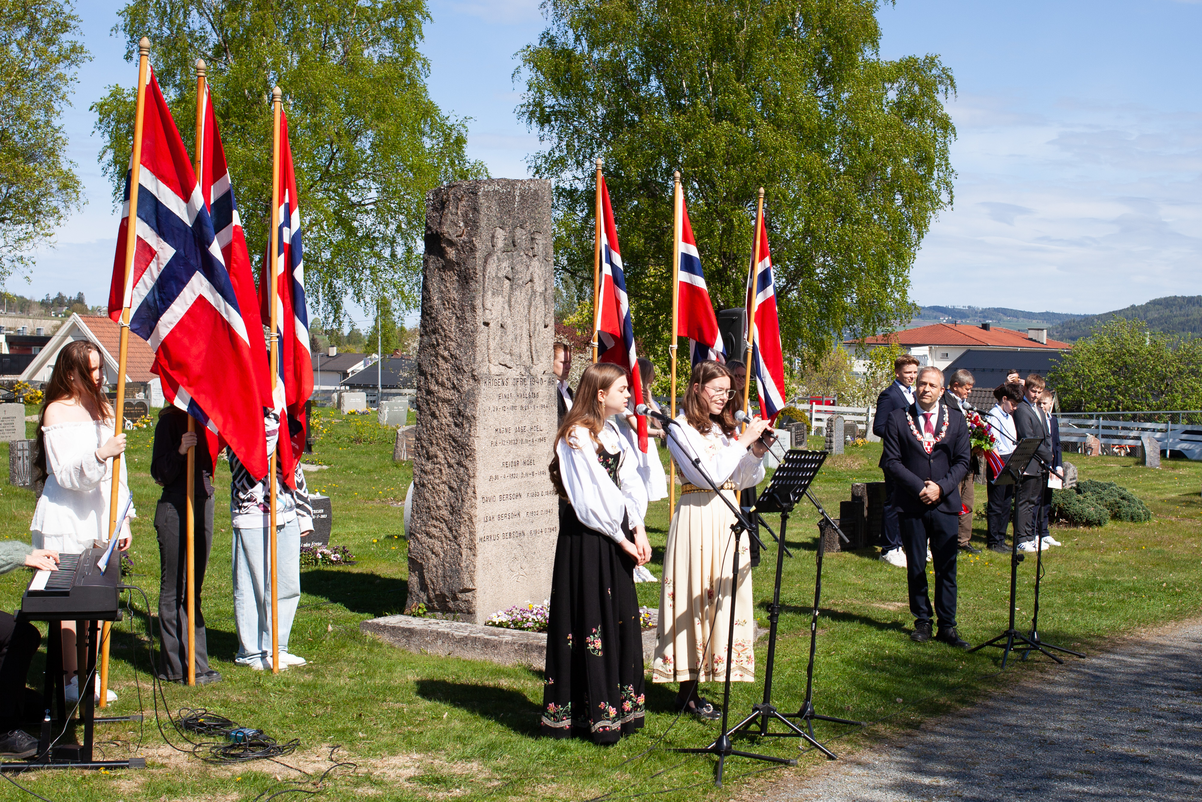 Bildet viser en flaggborg, en bauta, to bunadskledde som synger og en mann med ordførerkjede som ser på.