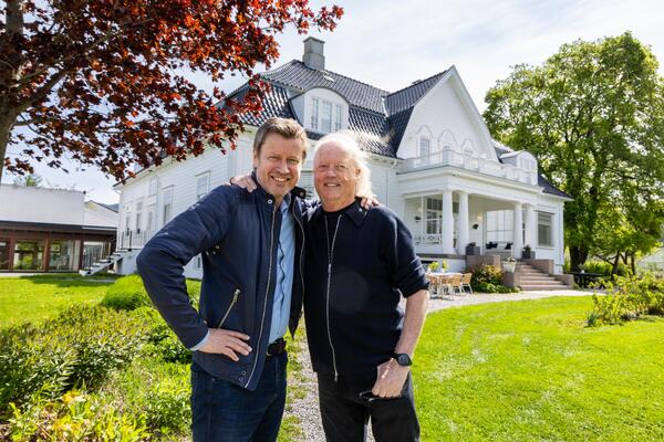 Bildet viser to storfornøyde personer som står ved siden av hverandre, med hendene opp på hverandres skulder. De står i en stor hage. Bak dem ser man en stor, ærverdig hvit bygning.