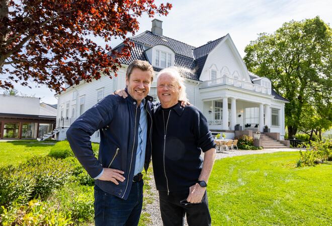Bildet viser to storfornøyde personer som står ved siden av hverandre, med hendene opp på hverandres skulder. De står i en stor hage. Bak dem ser man en stor, ærverdig hvit bygning.