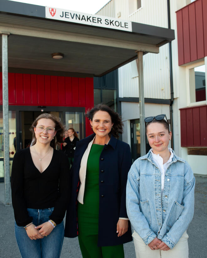 Bildet viser tre personer som står skulder ved skulder foran inngangen til et bygg. Over inngangen står det "Jevnaker skole".