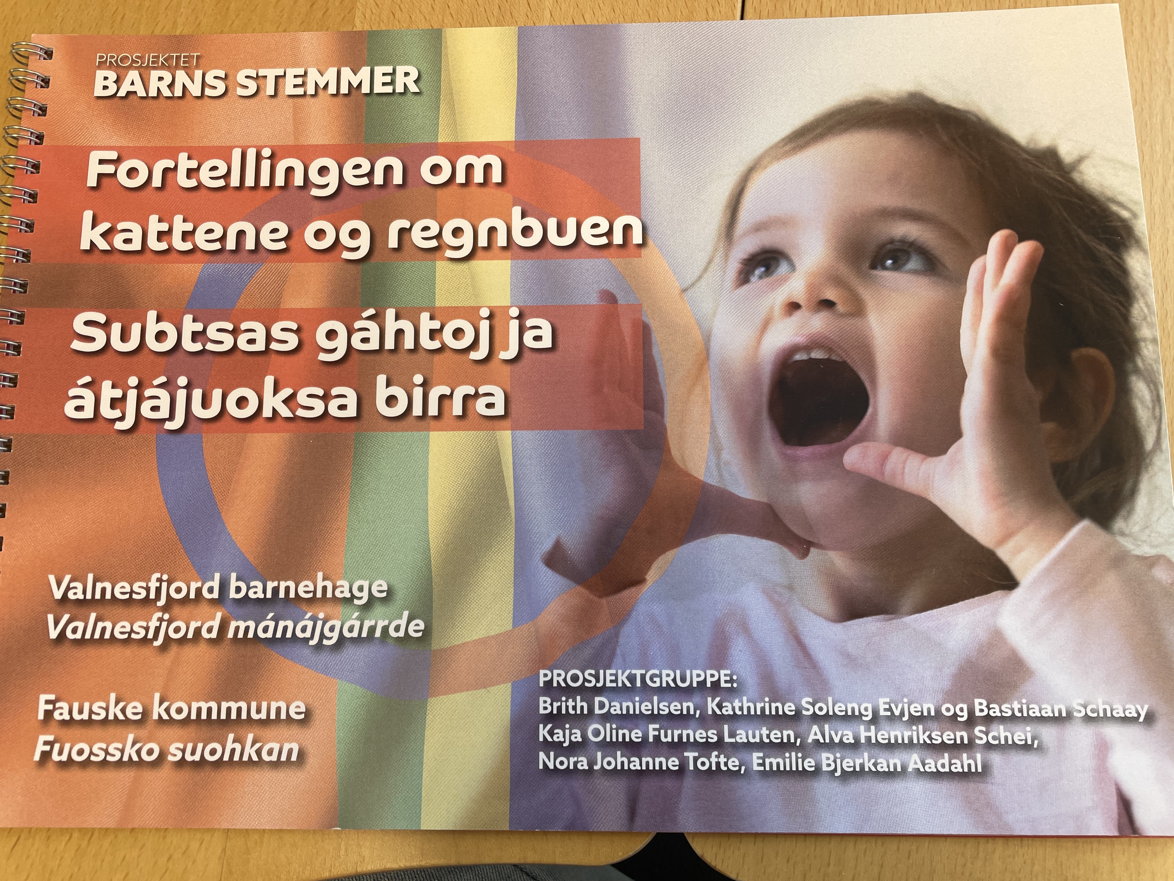 Hefte med fortellingen om kattene og regnbyen. Det er et samisk flagg og en jente med hendene på siden av en åpen munn