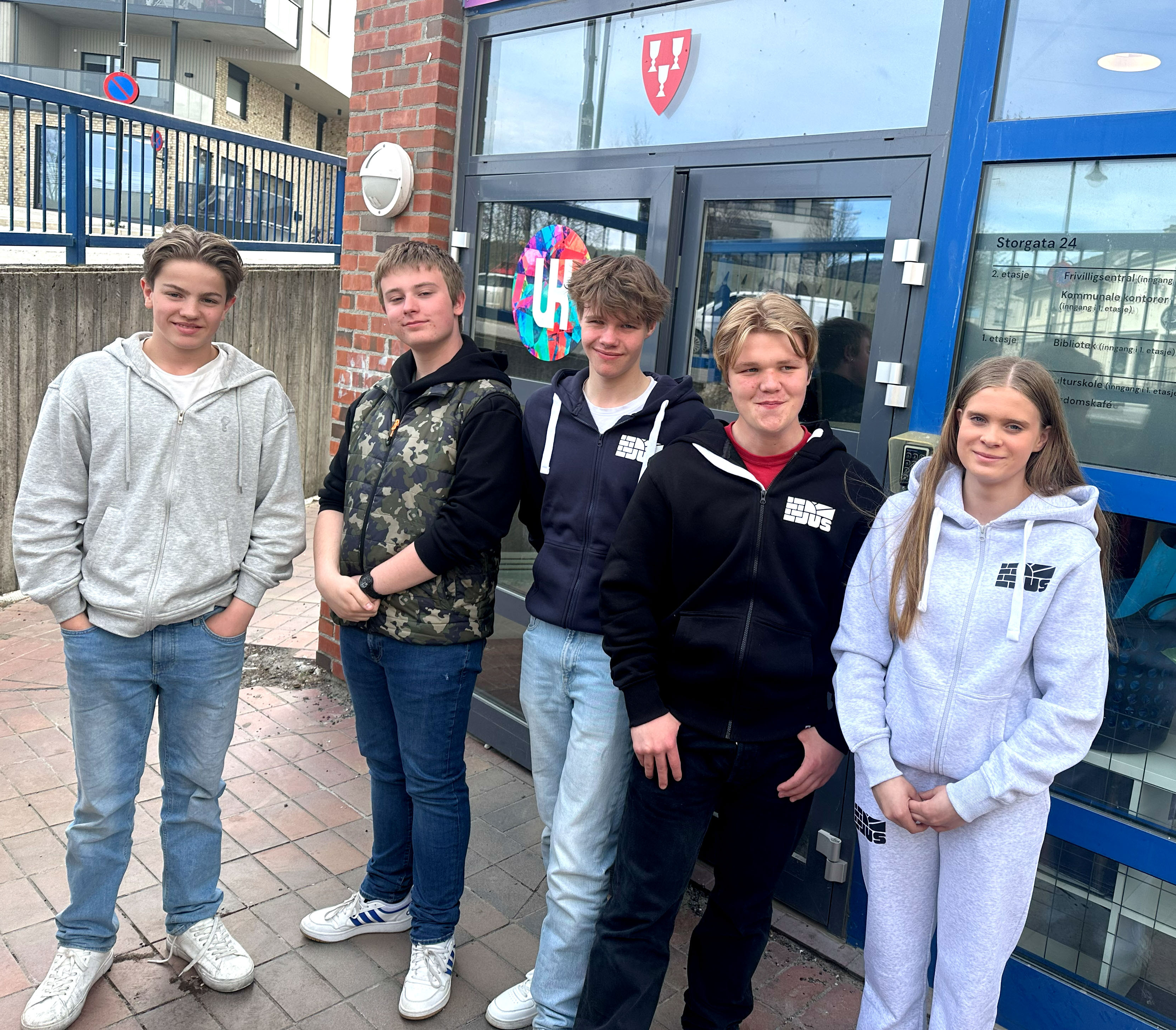 Bildet viser fem ungdommer som står ved siden av hverandre. De står foran en inngangsdør, hvor man ser både logoen til UK og kommunen.